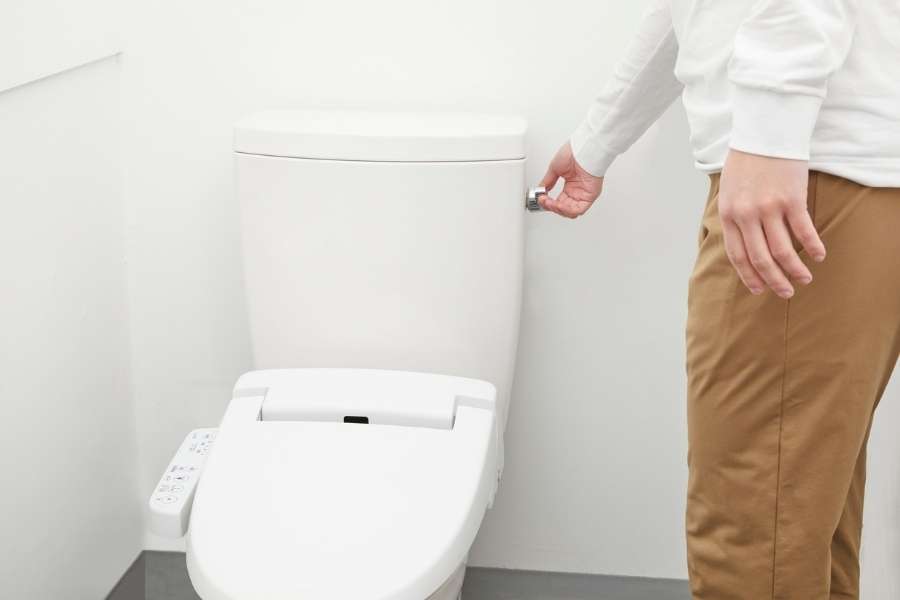 Une personne debout dans des toilettes modernes touche le bouton de commande latéral d'un réservoir blanc encastré, ce qui peut s'avérer nécessaire pour réparer une chasse d'eau.