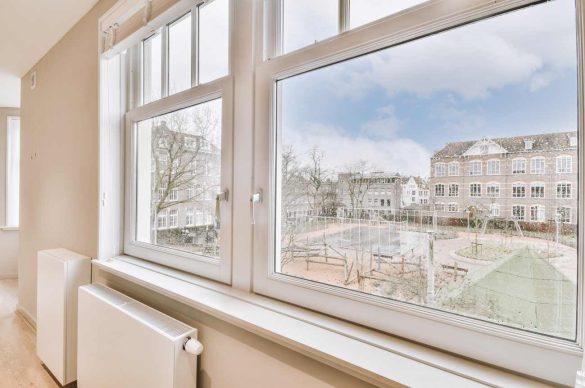 Vue d'intérieur sur des fenêtres PVC blanches donnant sur un immeuble ancien et un parc, illustrant la qualité de menuiserie idéale pour un achat réussi.