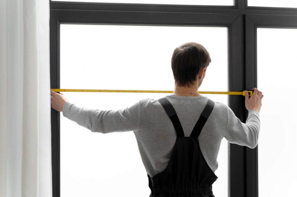 Un professionnel prend les mesures précises d'une grande fenêtre noire avec un ruban à mesurer jaune avant l'installation.